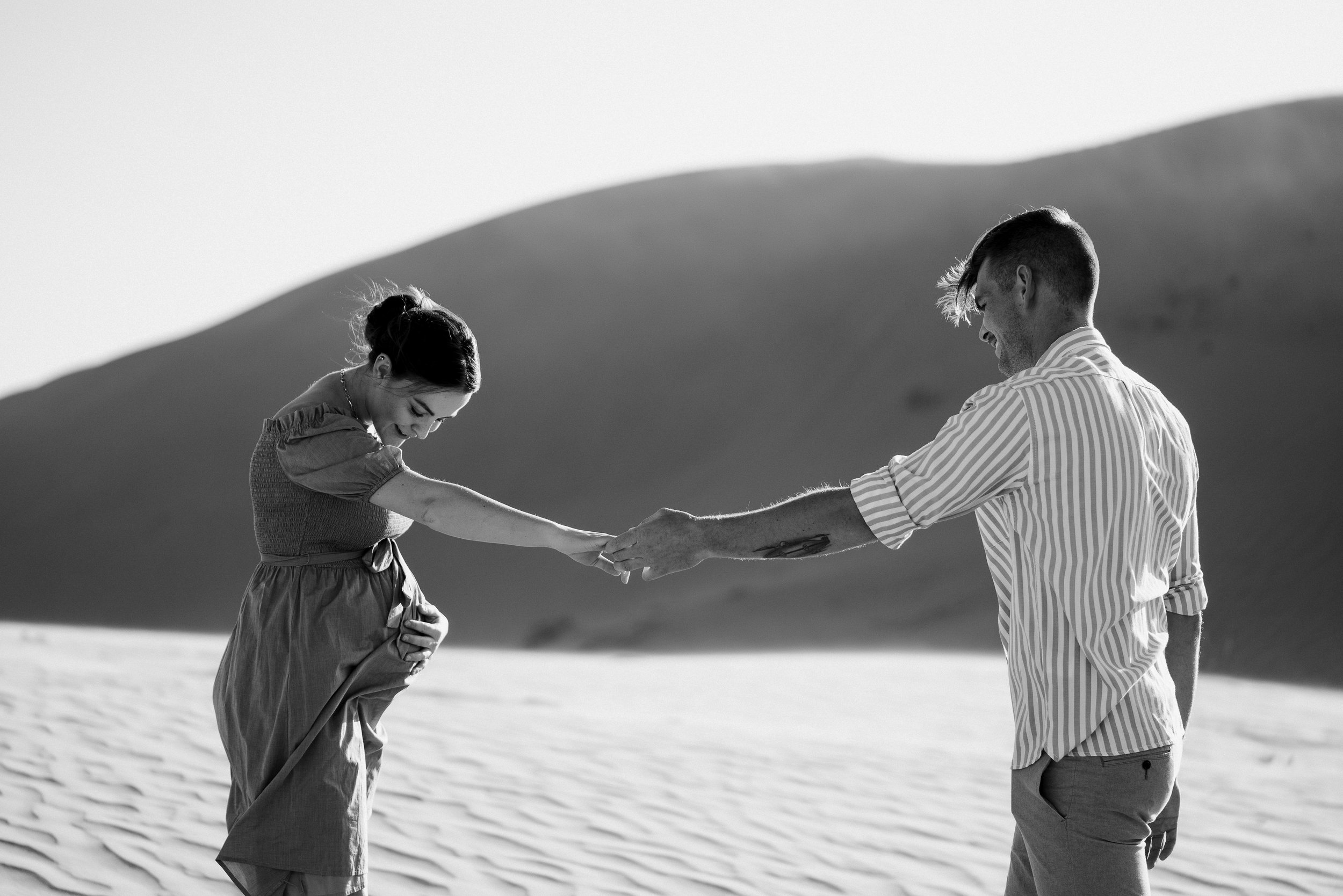 A couple in front of a large sand dune hold hands across some space, one partner is pregnant and is holding her belly and looking down. The other partner looks at her sweetly.