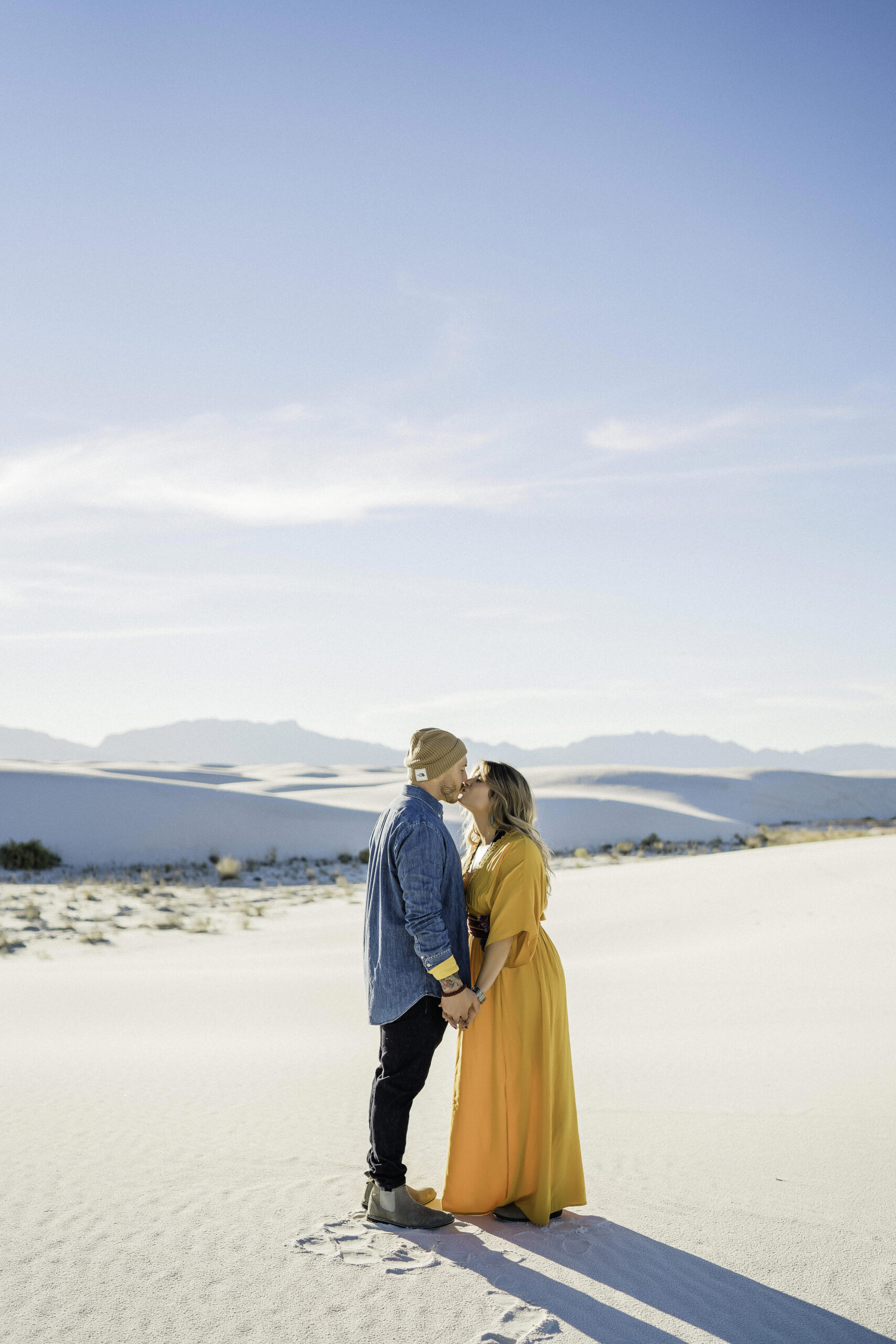 A couple kisses at white sands national park, in Alamogordo, New Mexico. She wears a yellow dress, he wears a denim button down and beanie.